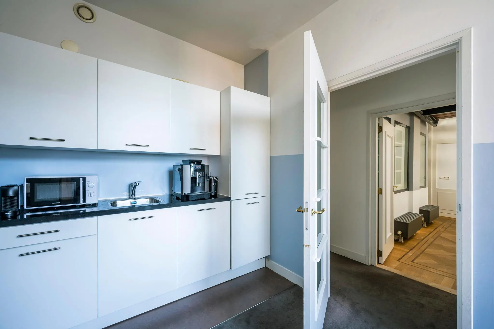 Moderne witte keuken aan de Prof. Tulpstraat met inbouwkasten, koffiezetapparaat en toegang tot een hal met houten vloer.